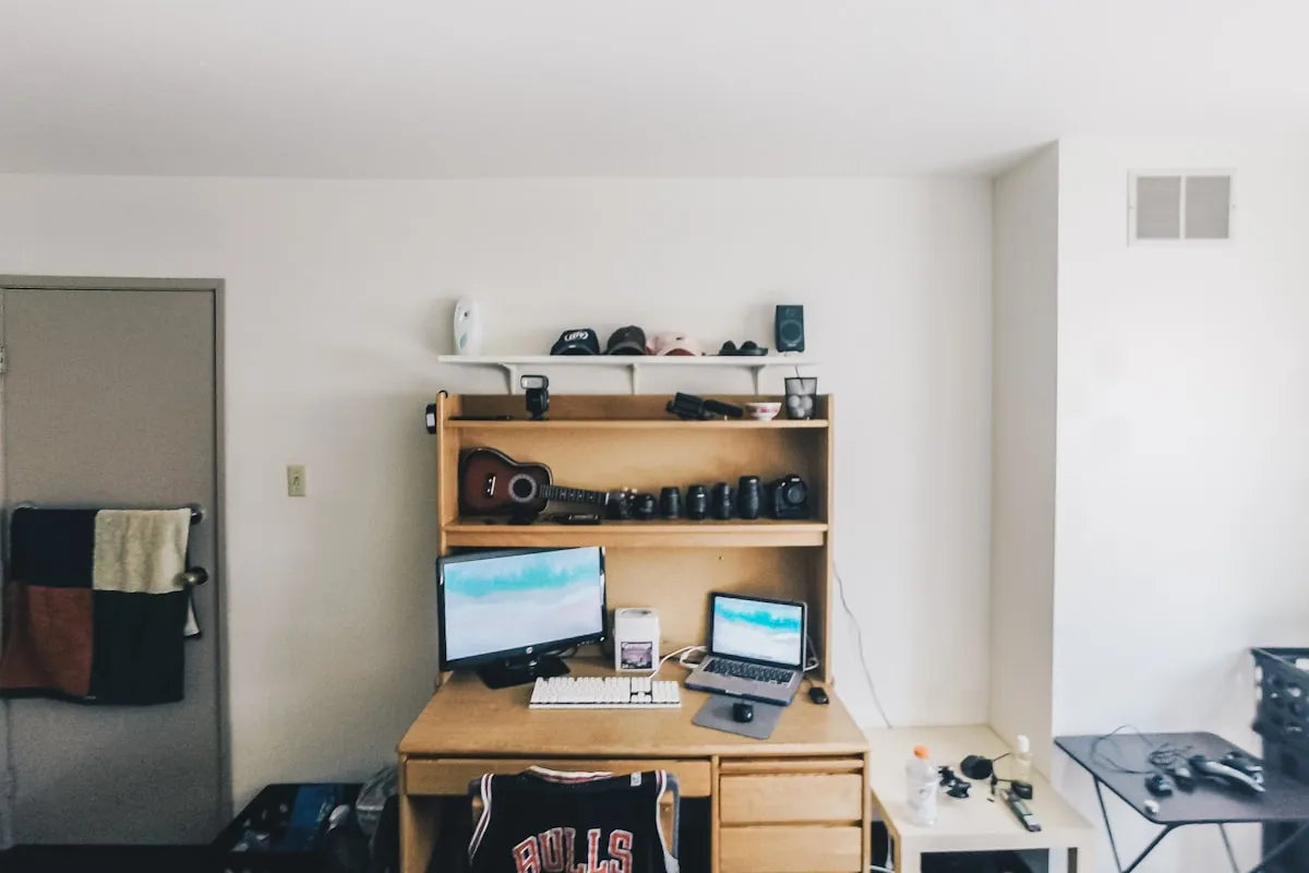 A dorm room desk with a wooden hutch shelf holding a guitar and camera gear, two computer monitors, and a basketball jersey draped over the chair