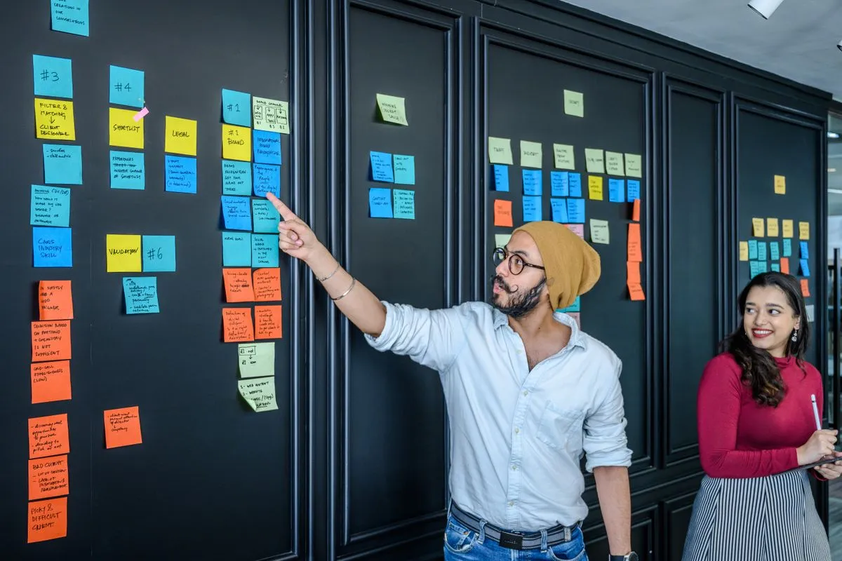 A man in a mustard beanie and glasses points at rows of blue, yellow and orange sticky notes on a black wall while a smiling colleague in a red top looks on
