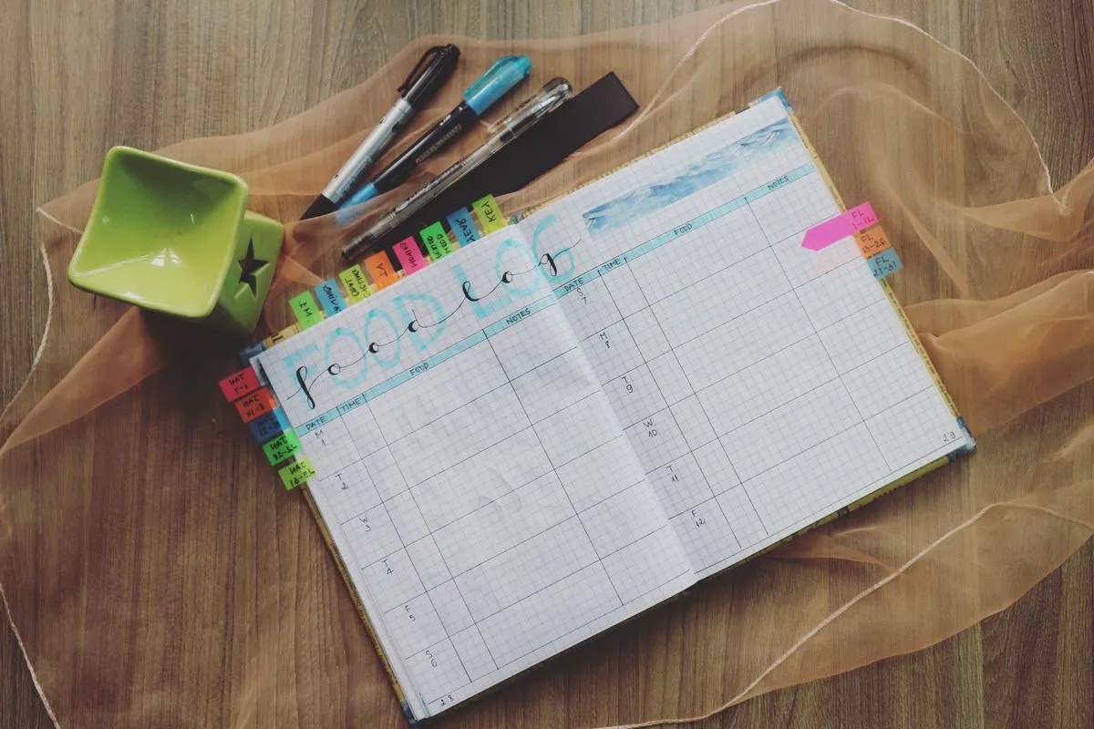An open hand-drawn food log notebook with date, time, food and notes columns and colorful page tabs, beside pens on a wooden table