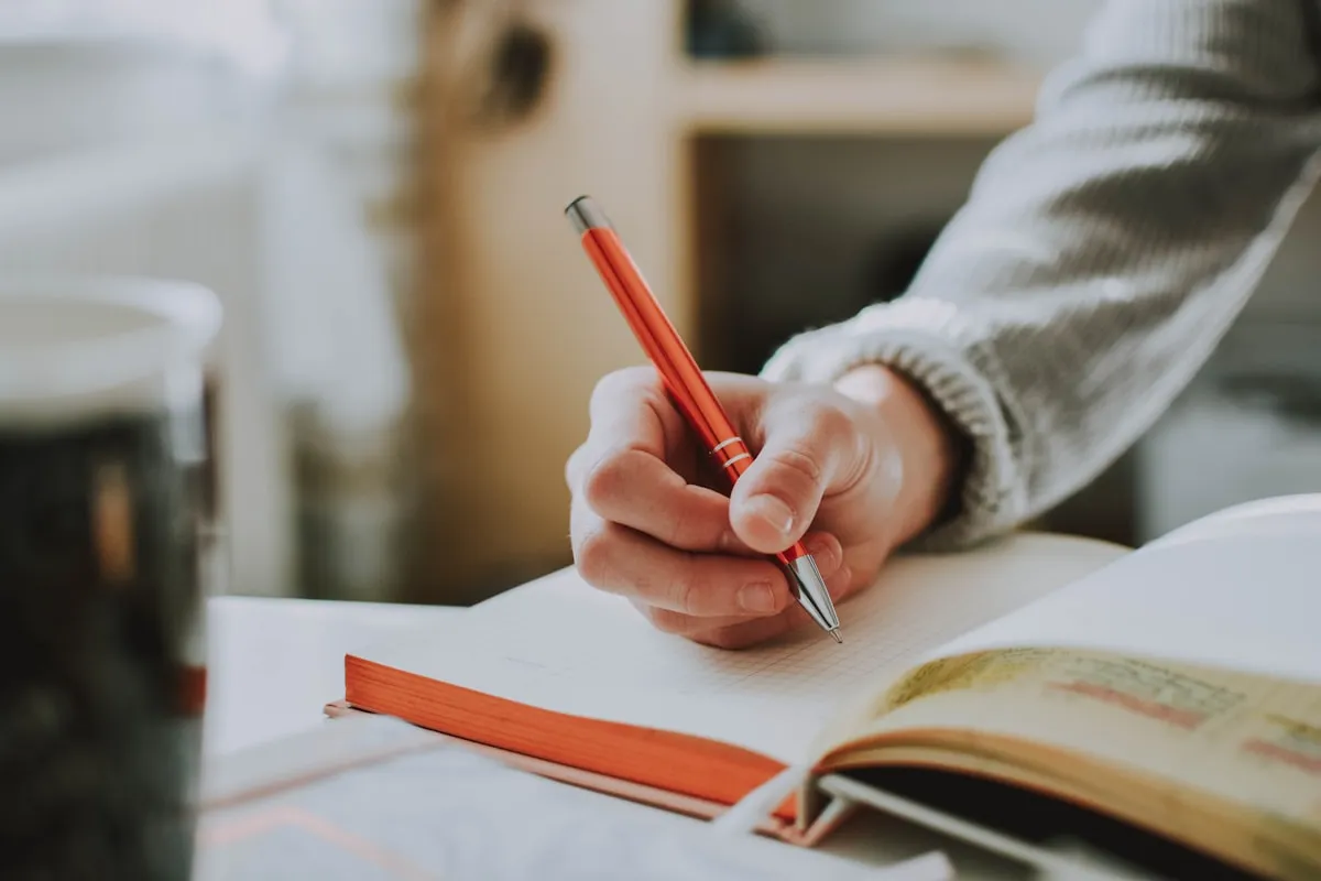 A hand in a grey knit sweater holds an orange pen over the blank grid pages of an open orange-edged notebook, beside a dark glass of coffee in warm window light