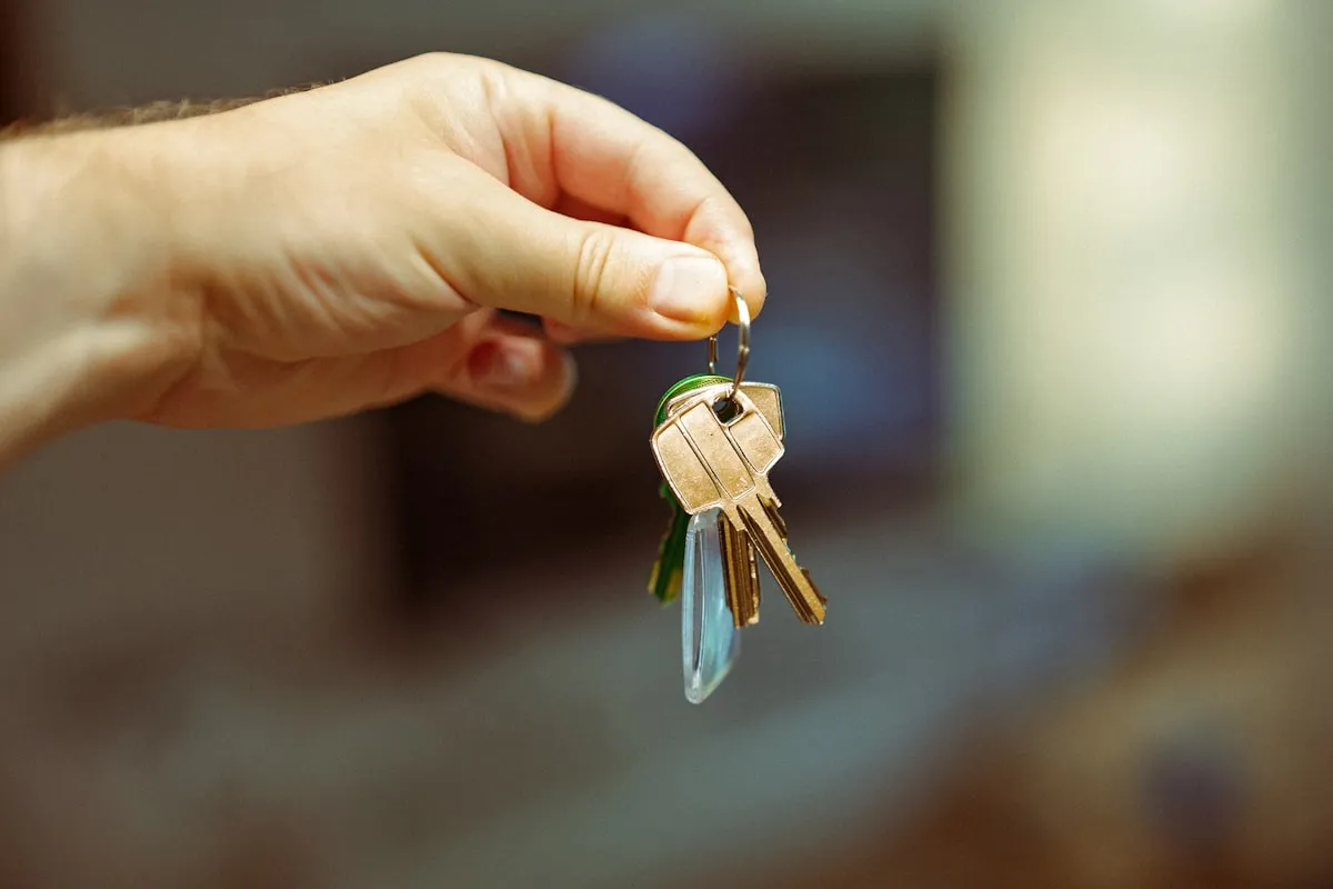 A hand holding out a small ring of brass door keys with a green rubber fob, against a softly blurred indoor background