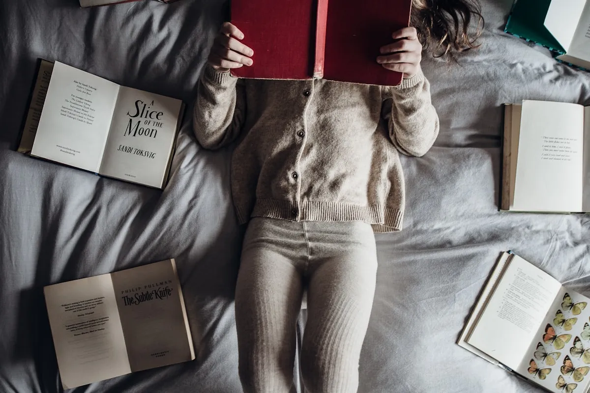 A child in a beige cardigan lying on a gray bed holding a red hardcover book over their face, surrounded by four open books laid on the bedsheets