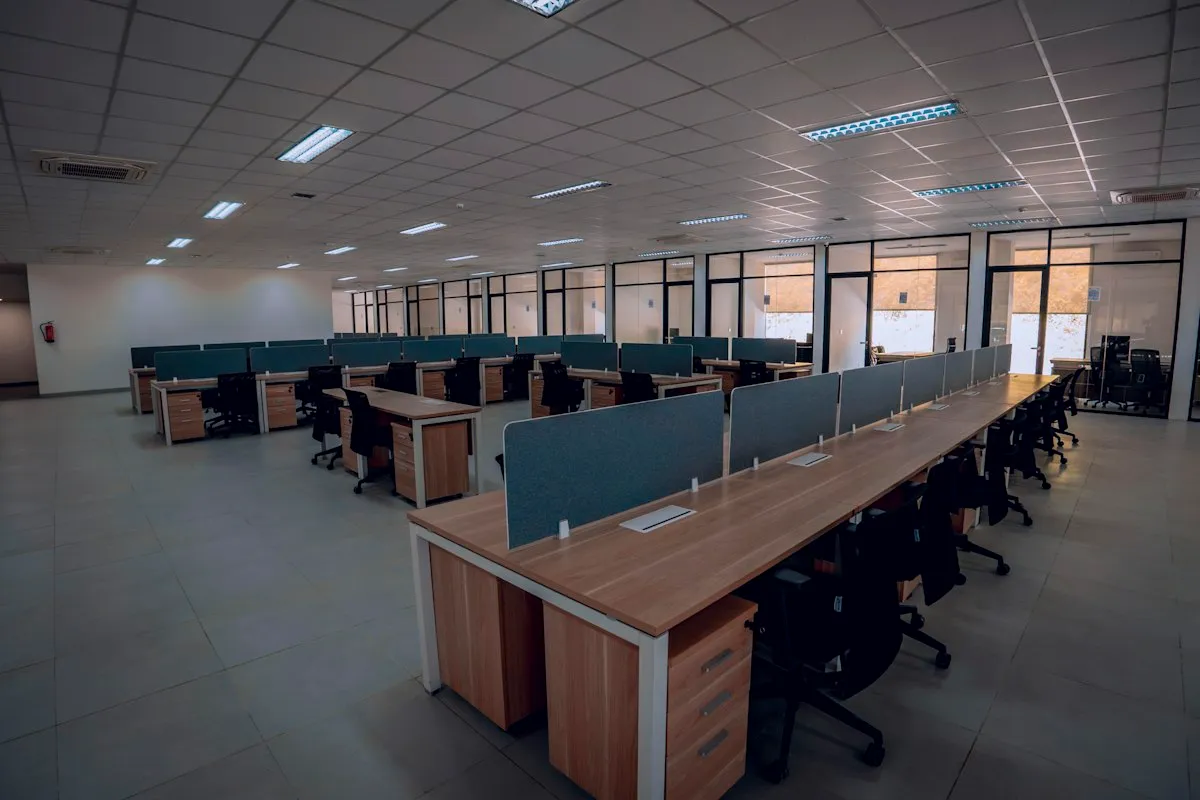 A large open-plan office with rows of empty wooden desks, gray fabric privacy panels and black swivel chairs under fluorescent ceiling lights, with no people present