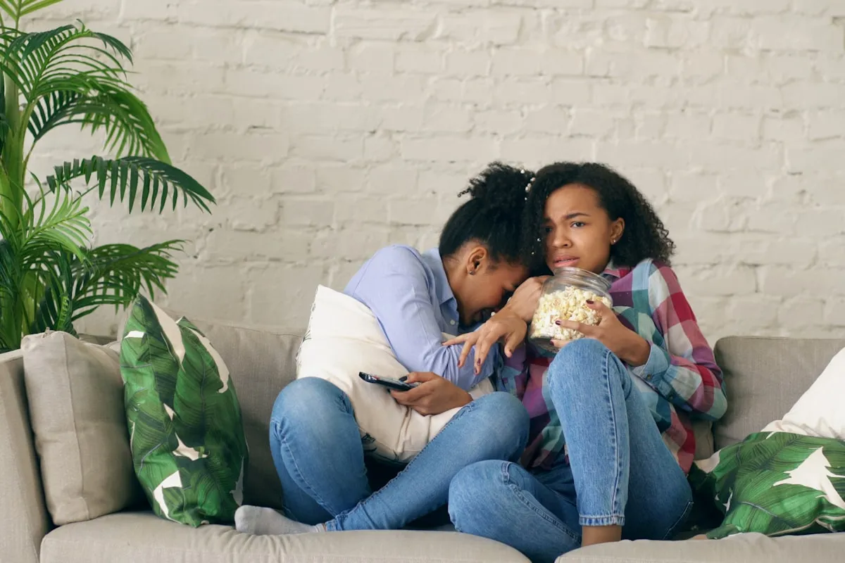 Two friends sitting close on a gray couch sharing a jar of popcorn, one hiding her face in the other's shoulder while reacting to something on the TV.