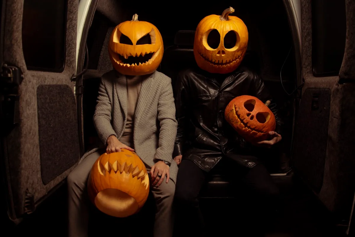 Two people wearing large carved jack-o'-lantern heads sitting side by side in the back of a dark van, each holding another carved pumpkin in their lap