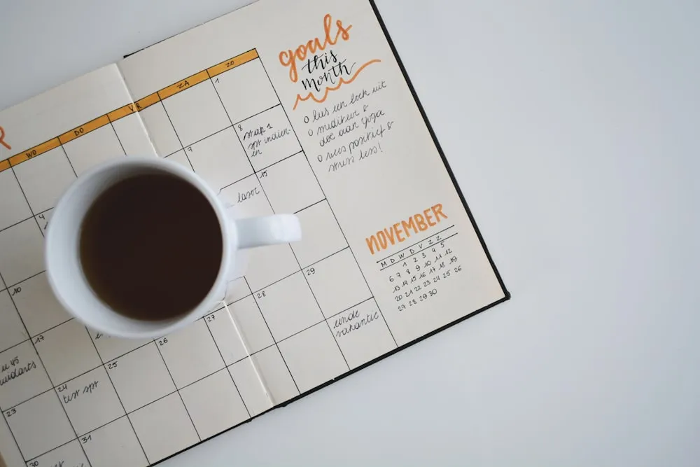A monthly planner open on a desk with a coffee cup, showing handwritten goals and a calendar grid.