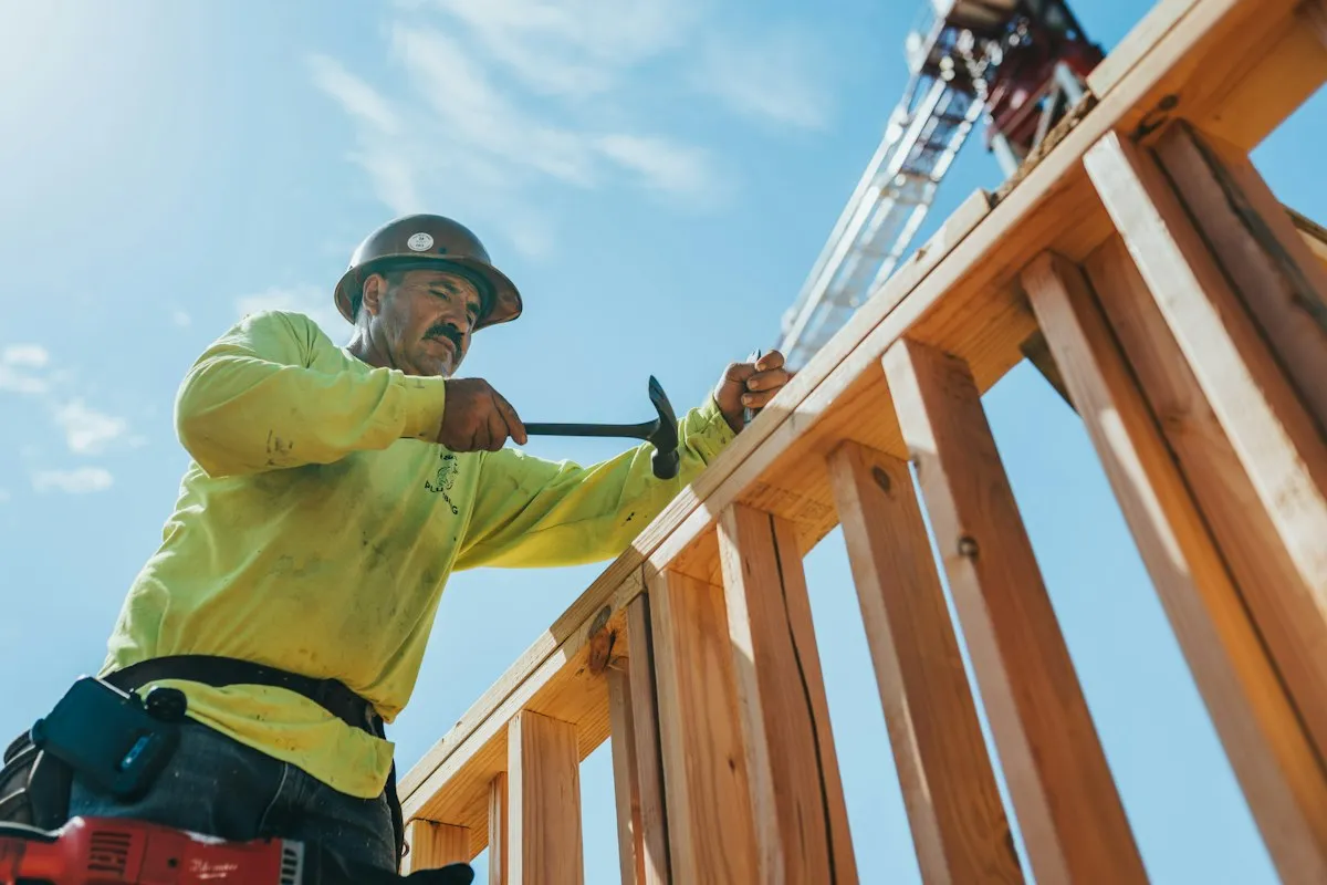 Construction worker in a hard hat hammering timber wall framing high up, with a blue sky and a crane behind him