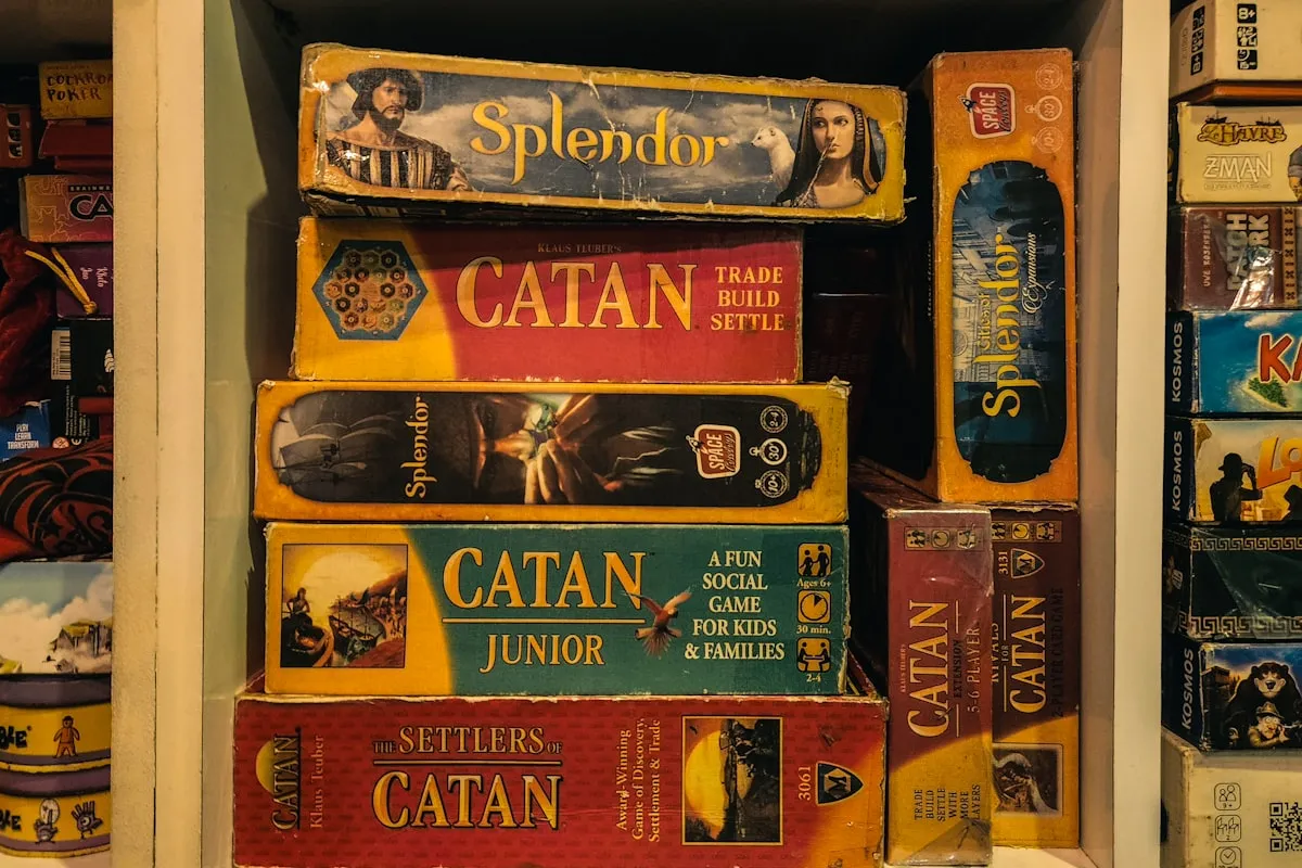 Stacked board game boxes including Splendor and Catan filling a shelf, with more game boxes shelved upright on either side
