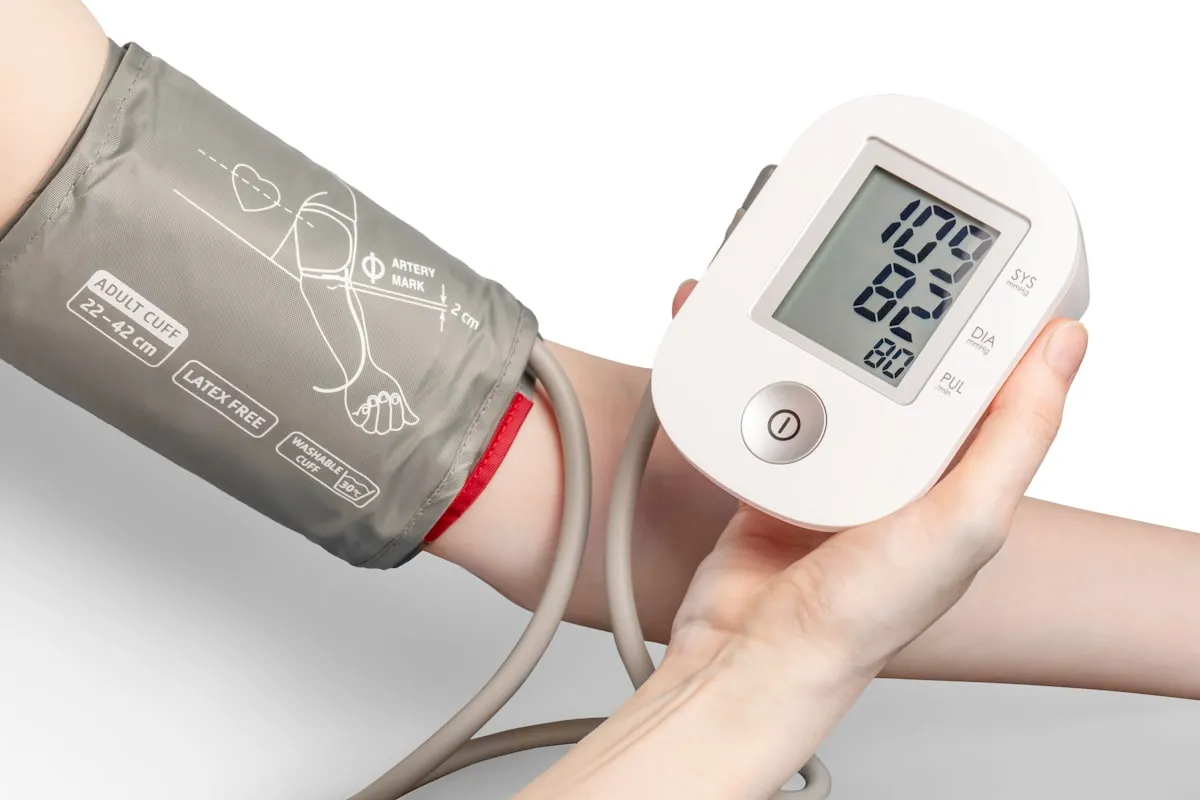 A bare arm in a gray blood pressure cuff while a hand holds a white digital monitor reading 109 over 82 with a pulse of 80, against a plain white background