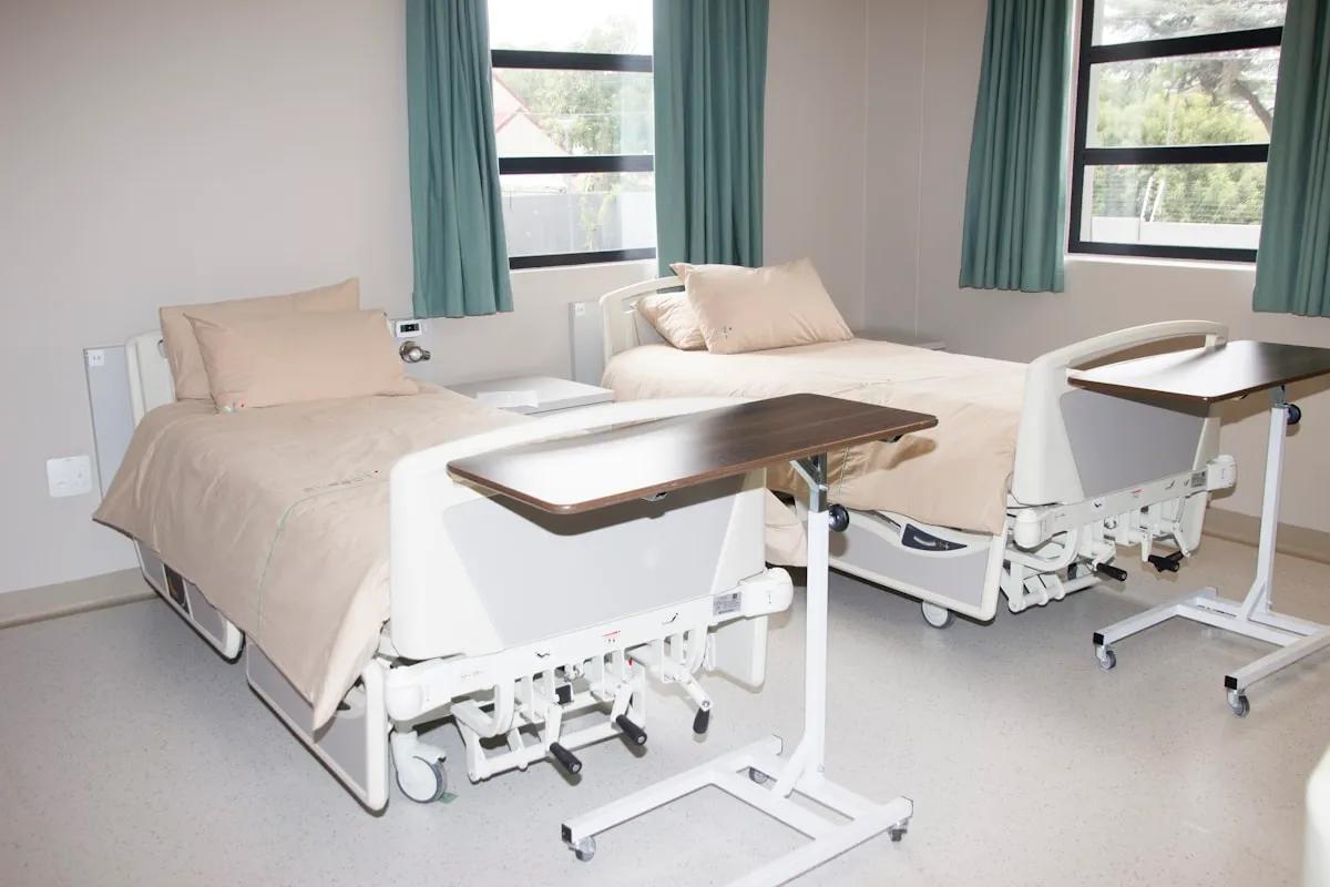 Two empty hospital beds with beige blankets and pillows in a bright shared ward, wheeled overbed tray tables pulled across them, green curtains drawn back from two windows