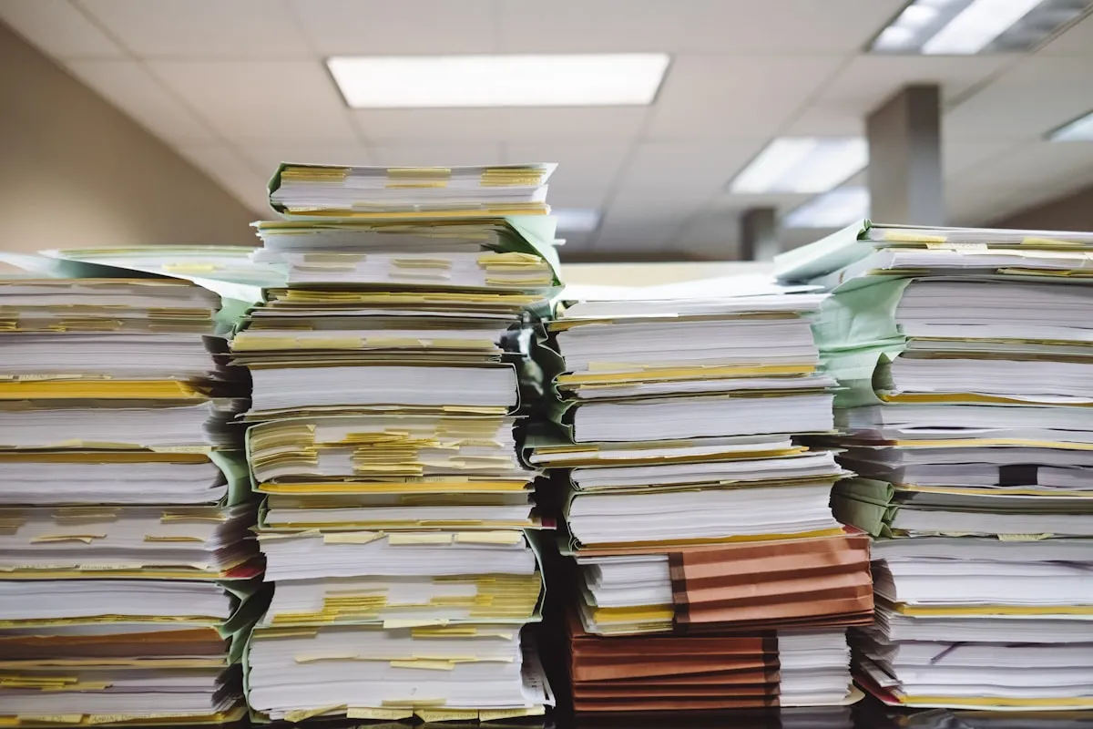 Tall stacks of white and yellow paper documents in manila and green file folders piled on a desk in a fluorescent-lit office, with dozens of yellow tabs marking pages