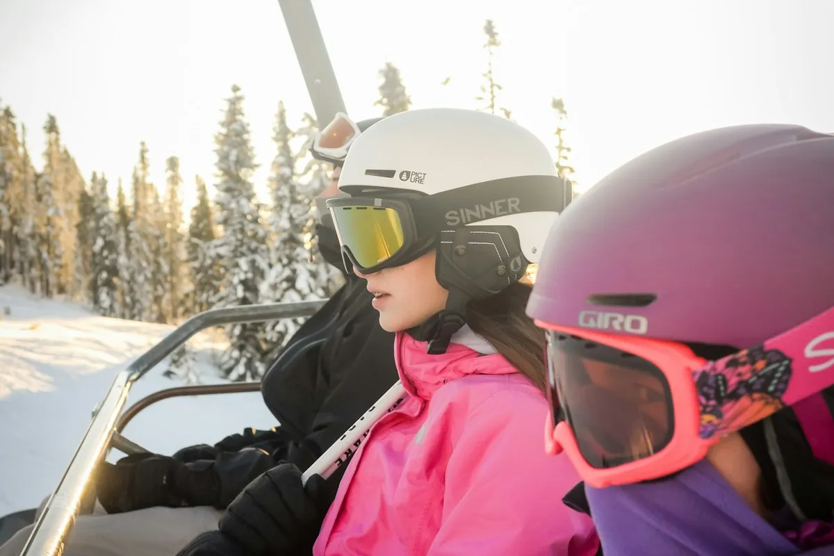 A family on a chairlift in winter sun, a teen in a white helmet and pink jacket beside a rider in a purple helmet and pink goggles, with snow-covered pines behind