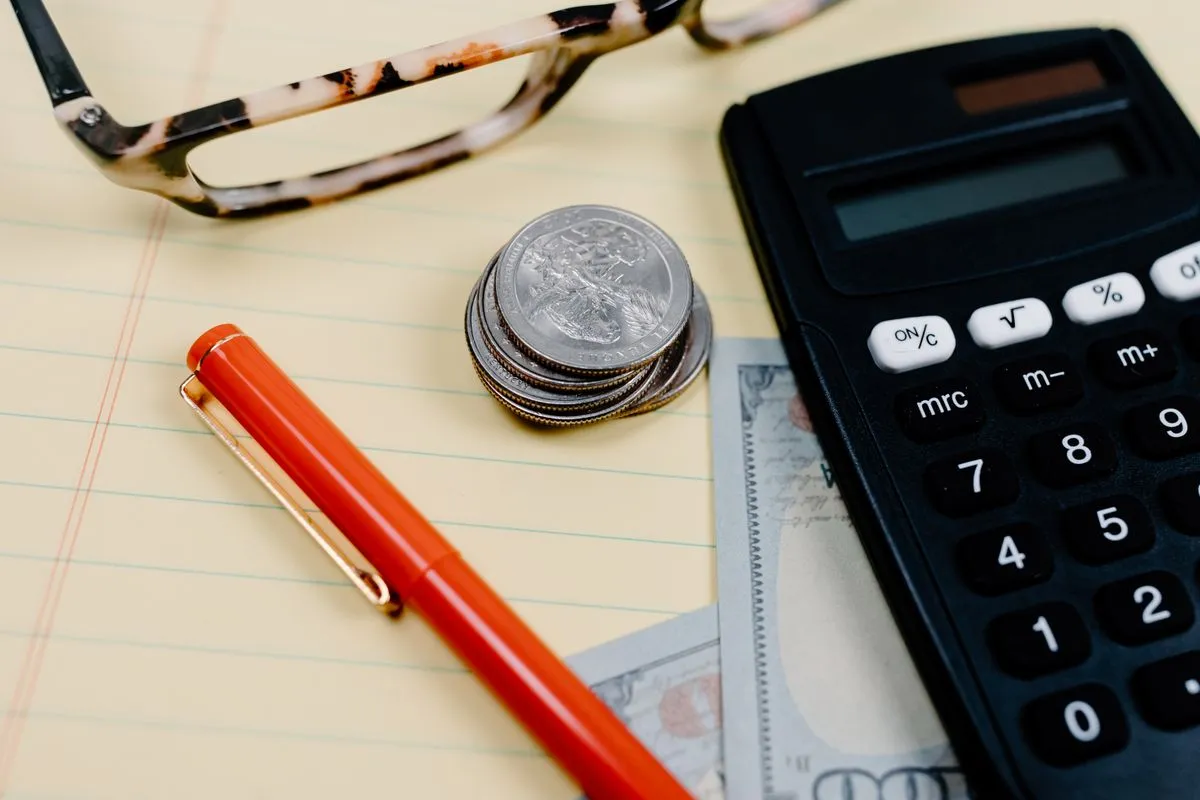 A small stack of coins on a yellow legal pad beside an orange pen, a black desk calculator, two banknotes and tortoiseshell reading glasses