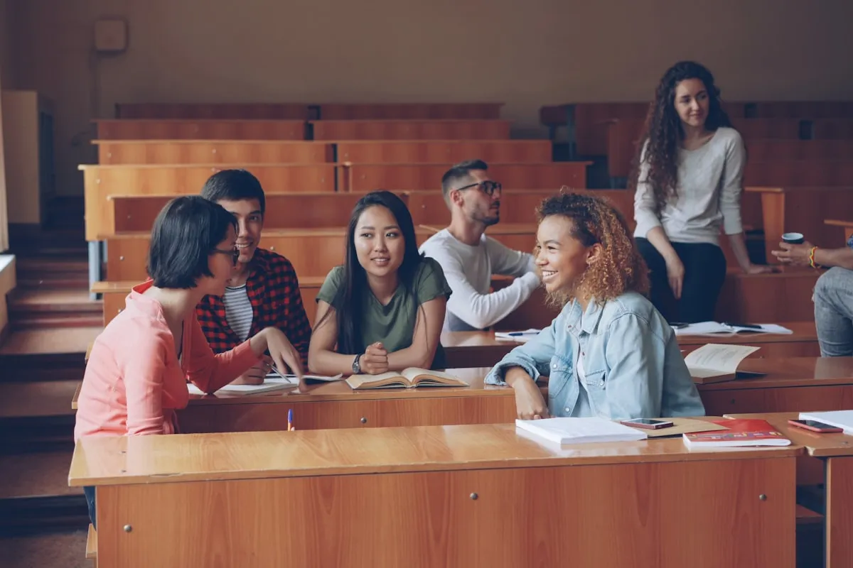 College students at tiered wooden desks in a lecture hall, several talking and smiling over open textbooks while another leans against a desk behind them