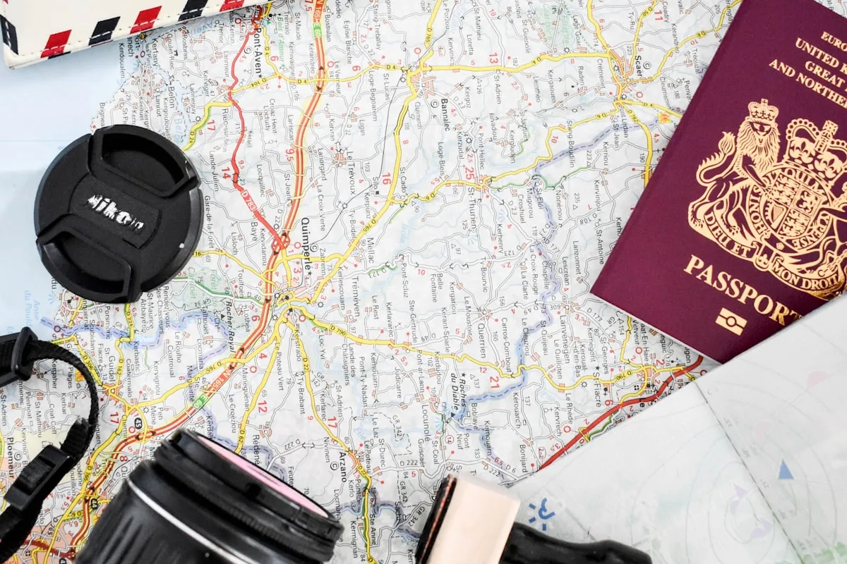 A detailed road map covered with a maroon UK passport, a camera with a Nikon lens cap, and folded travel documents arranged on a table