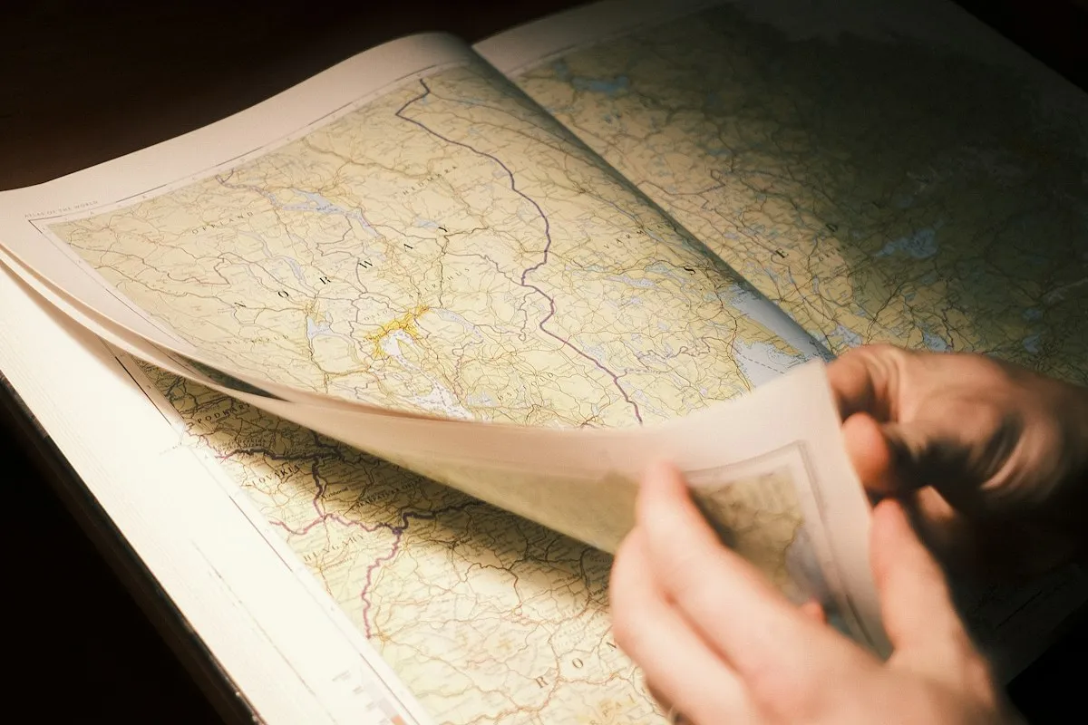 A hand turning the page of a large printed atlas open to a map of Norway and Sweden, lit by warm lamplight against a dark tabletop