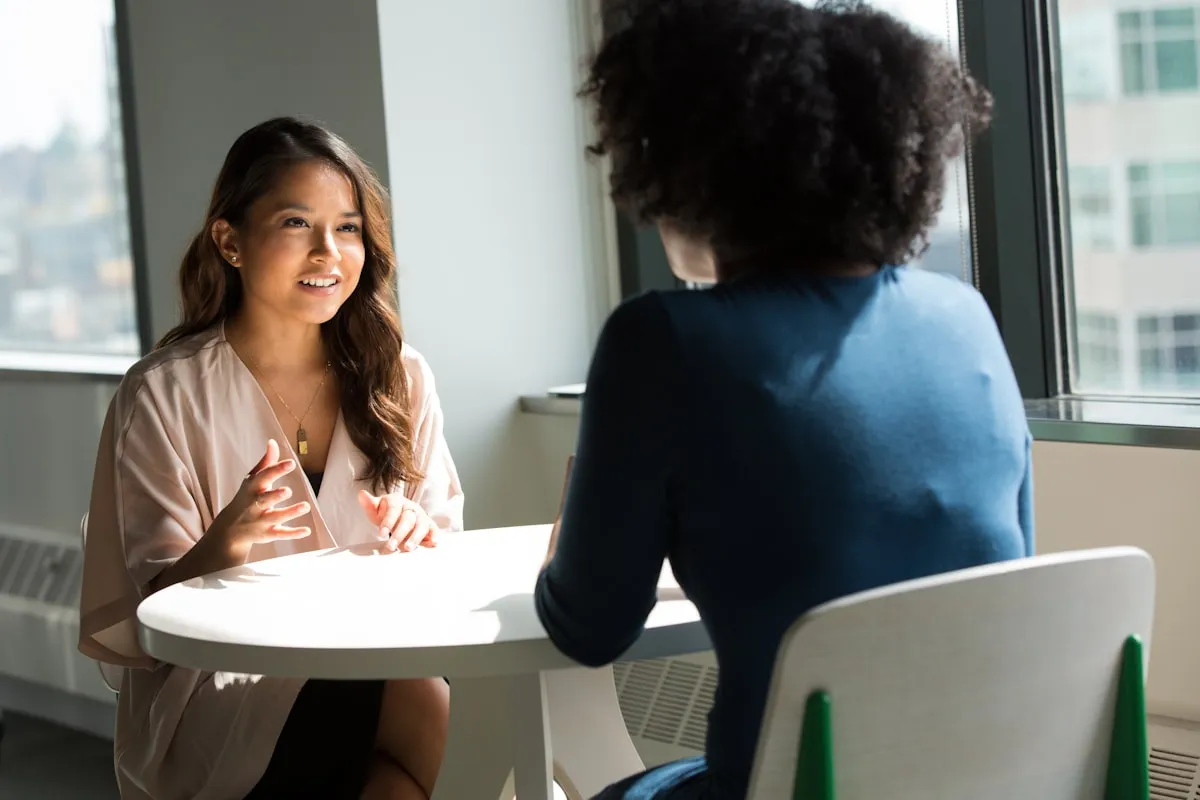 Two women talking across a small round table by a window in a bright office, one gesturing mid-sentence while the other listens with her back to the camera