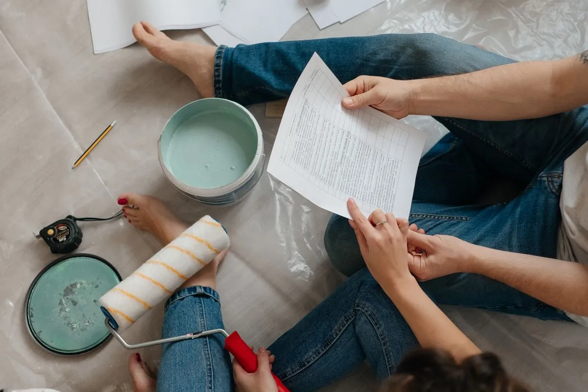Overhead view of two people sitting on a plastic drop cloth in jeans, holding a printed itemized sheet, beside an open can of mint-green paint, a paint roller and a tape measure
