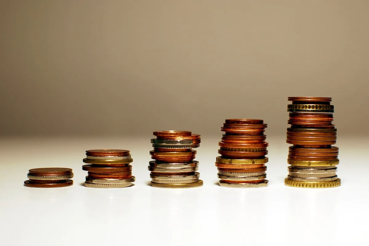 Five stacks of assorted copper, brass and silver coins on a white surface, arranged left to right from a two-coin stack up to a stack about twelve coins high