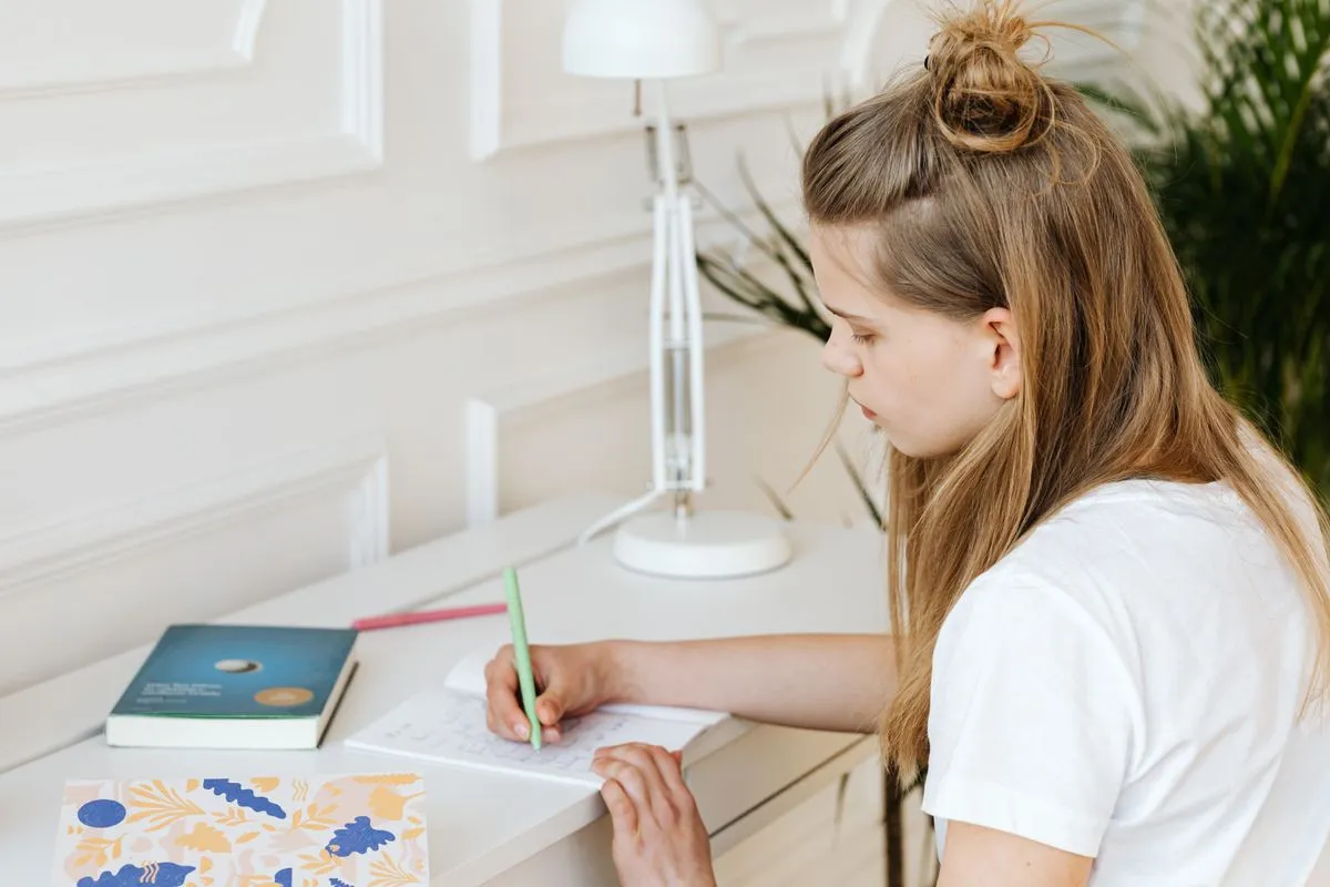 A teenage girl with her hair in a half-bun writes with a green pen in an open notebook at a white desk beside a white lamp, a book and a patterned folder