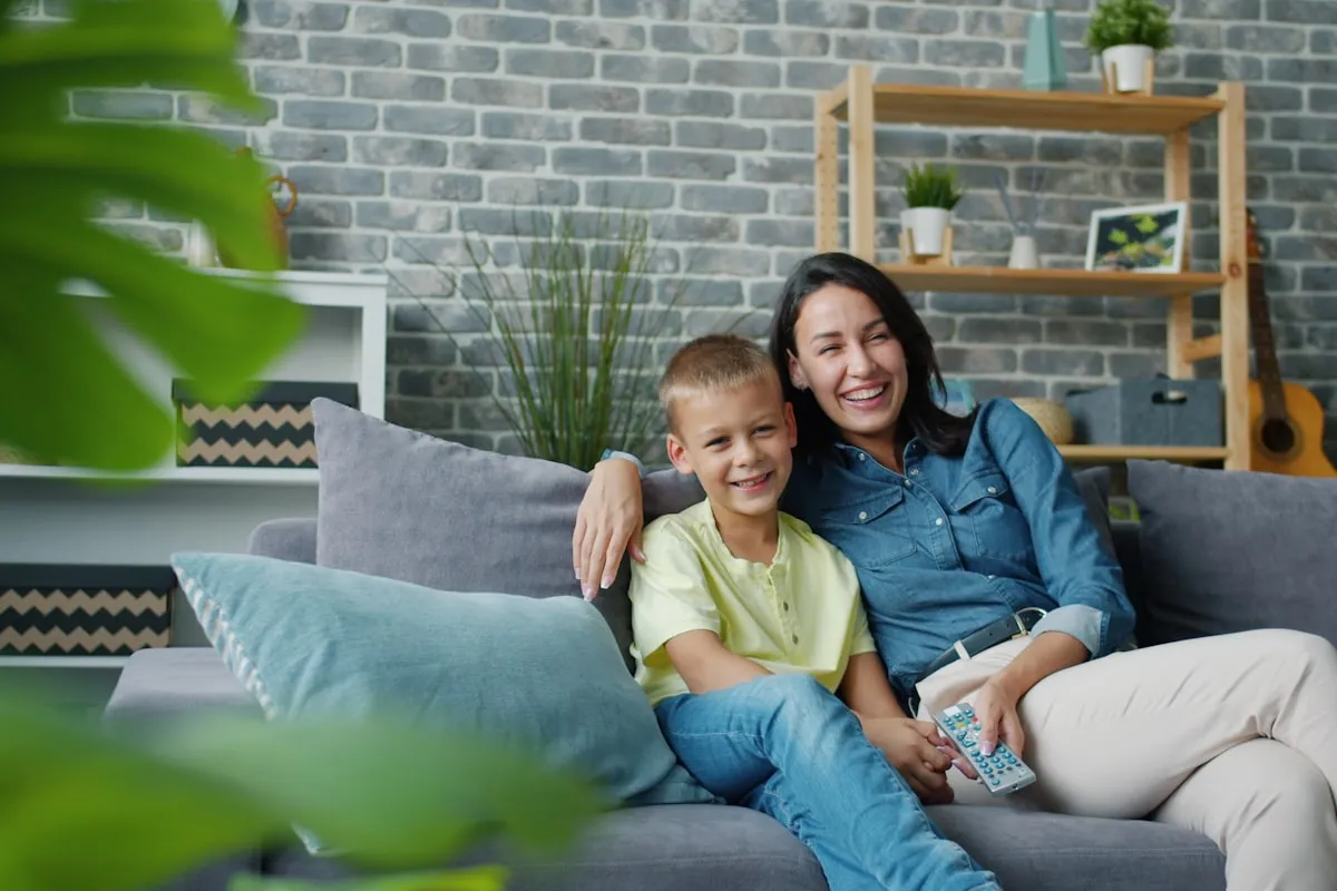 A smiling woman and a young boy sitting together on a gray sofa in a living room with a brick wall, shelves and a guitar behind them, the woman holding a TV remote