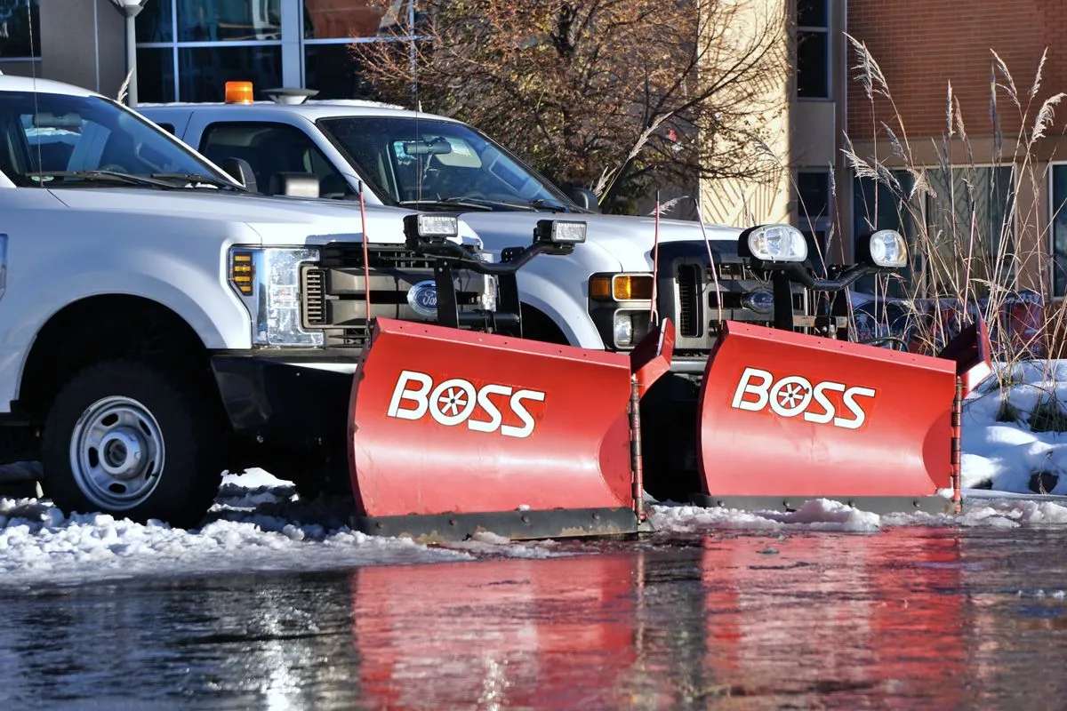 Two white pickup trucks parked side by side with red V-shaped snow plow blades mounted on their fronts, on a wet, slushy lot beside a brick building
