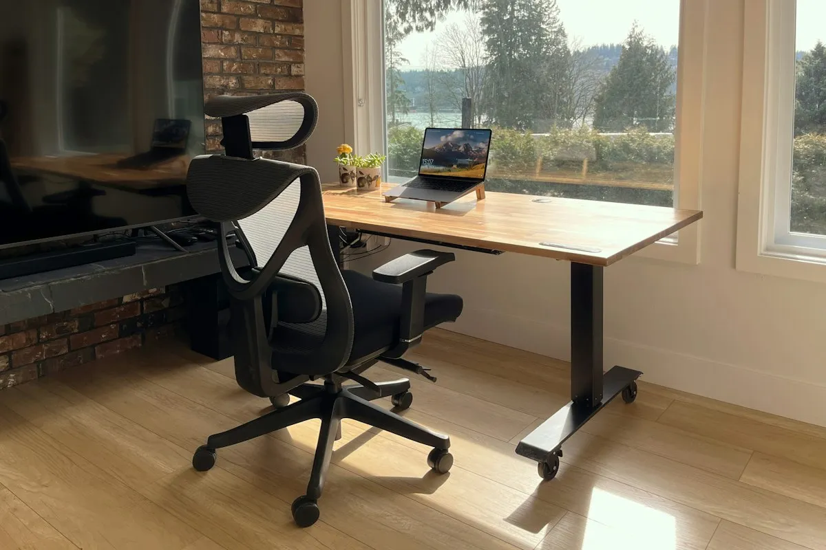 A black mesh ergonomic office chair beside a height-adjustable butcher-block desk with a laptop on a stand, by a sunny window