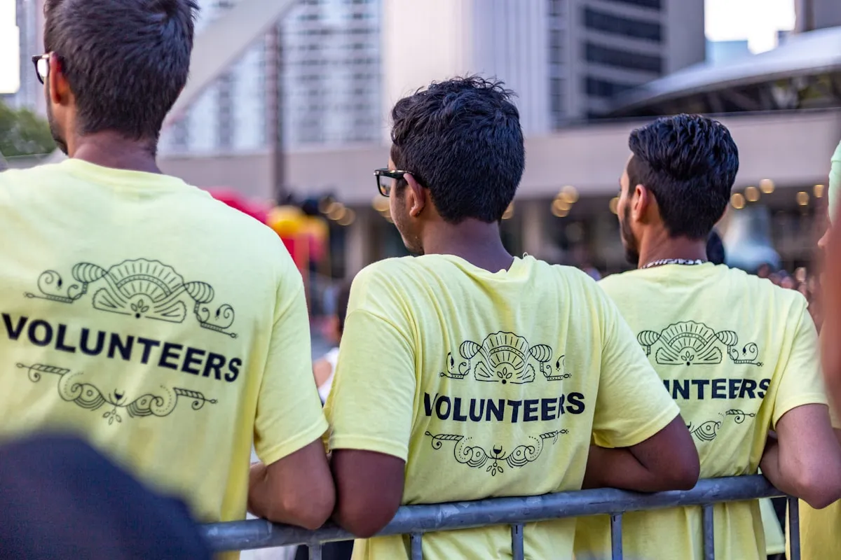 Three volunteers in pale yellow shirts printed with the word VOLUNTEERS, seen from behind, leaning on a metal crowd barrier at an outdoor city event