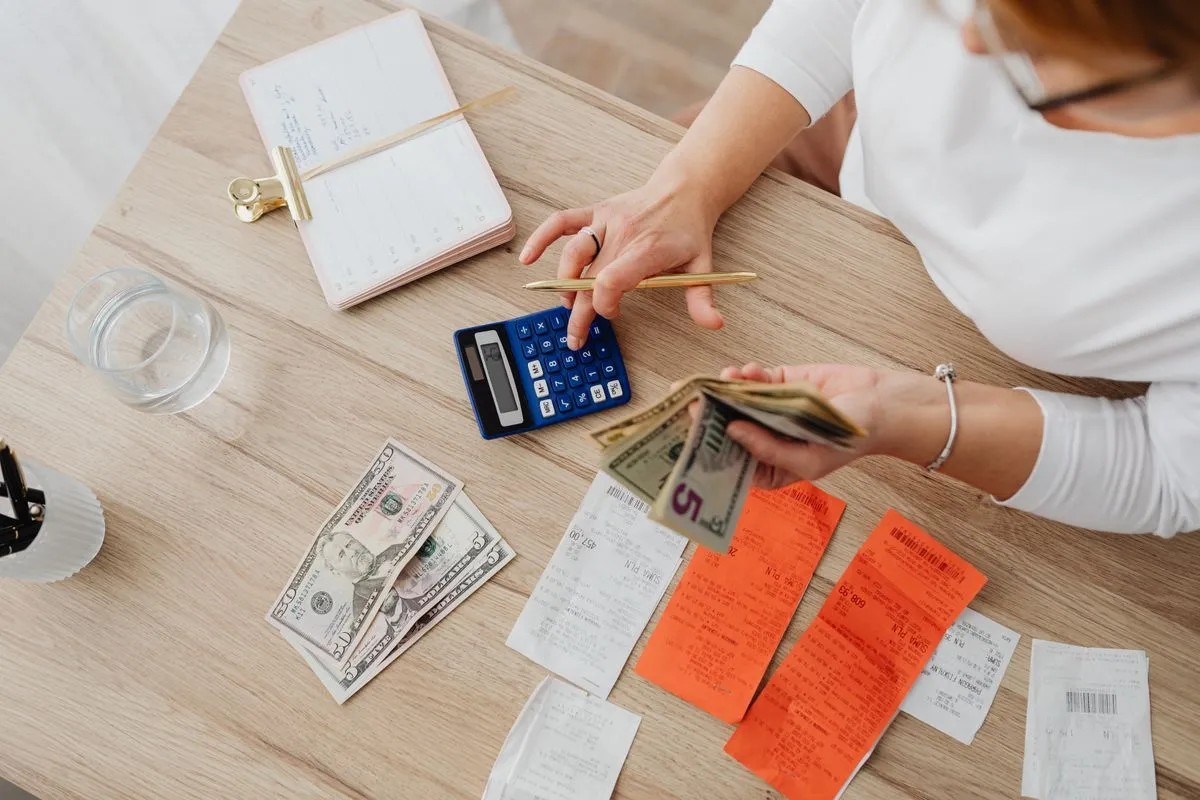 Hands holding cash and a pen over a calculator, with receipts and an open notebook on a wooden table