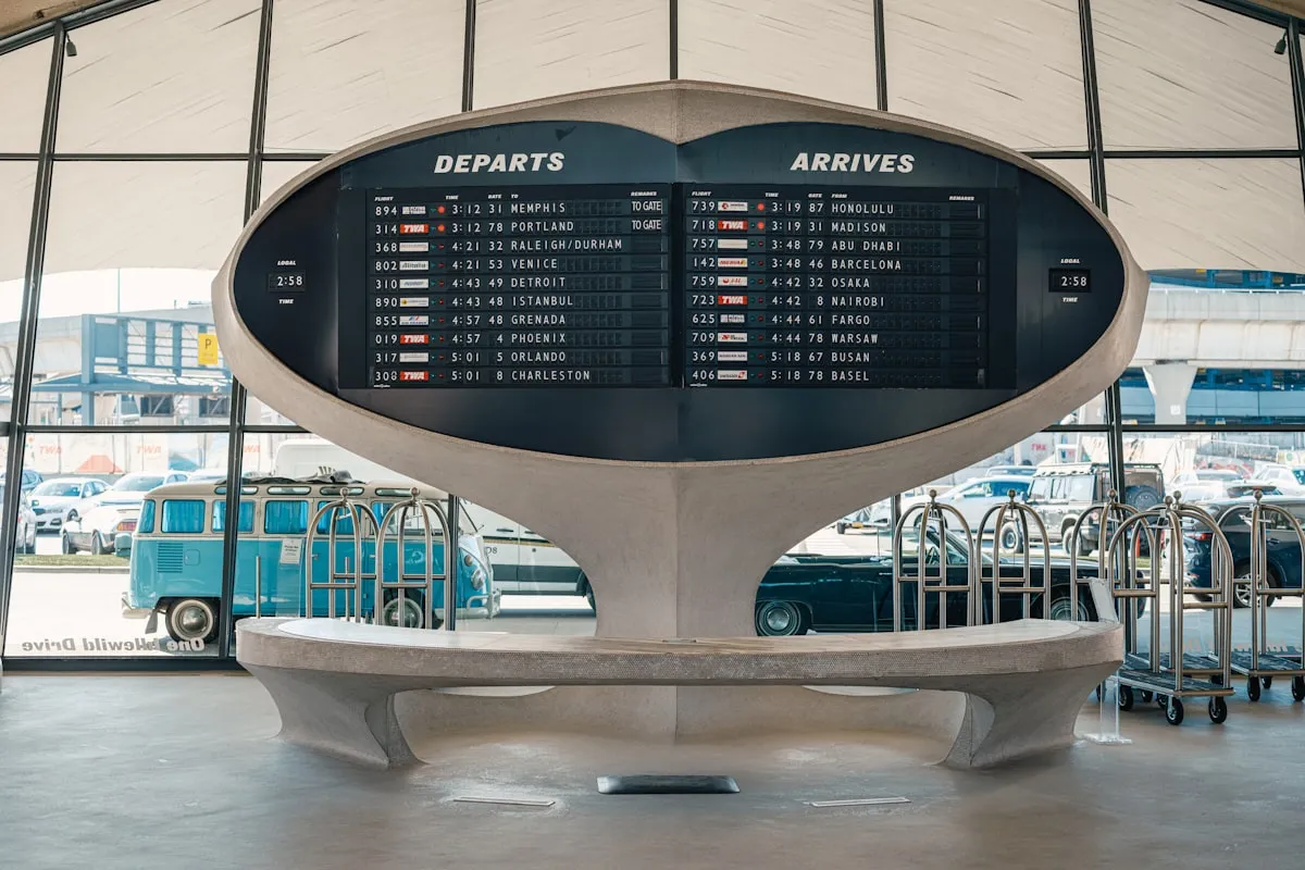 A retro split-flap airport board headed DEPARTS and ARRIVES, mounted on a curved concrete pedestal in a terminal, with brass luggage carts and a vintage van visible through the windows behind it