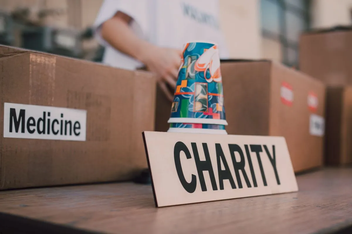 A cardboard box labeled Medicine and a wooden sign reading Charity on a table, a stack of patterned paper cups behind the sign, with a volunteer in a white shirt sorting donation boxes in the background