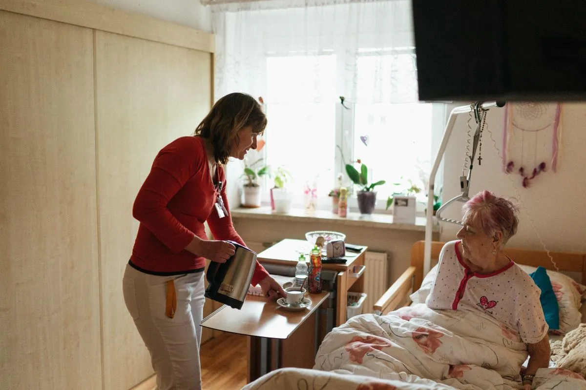 A caregiver with an ID badge pours hot water from a kettle into a teacup for an older woman sitting up in bed in a bright room with plants on the windowsill