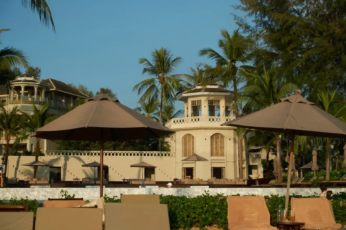 A cream-colored beachfront resort building with an octagonal tower, surrounded by tall palm trees, dark canvas umbrellas, and empty poolside loungers under a clear blue sky