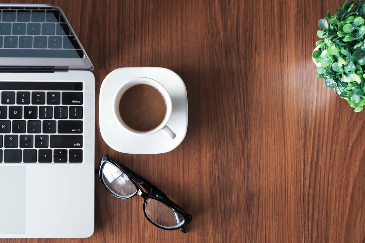 Overhead view of an open laptop, a cup of black coffee on a white saucer, black eyeglasses, and a small green plant on a polished wooden desk