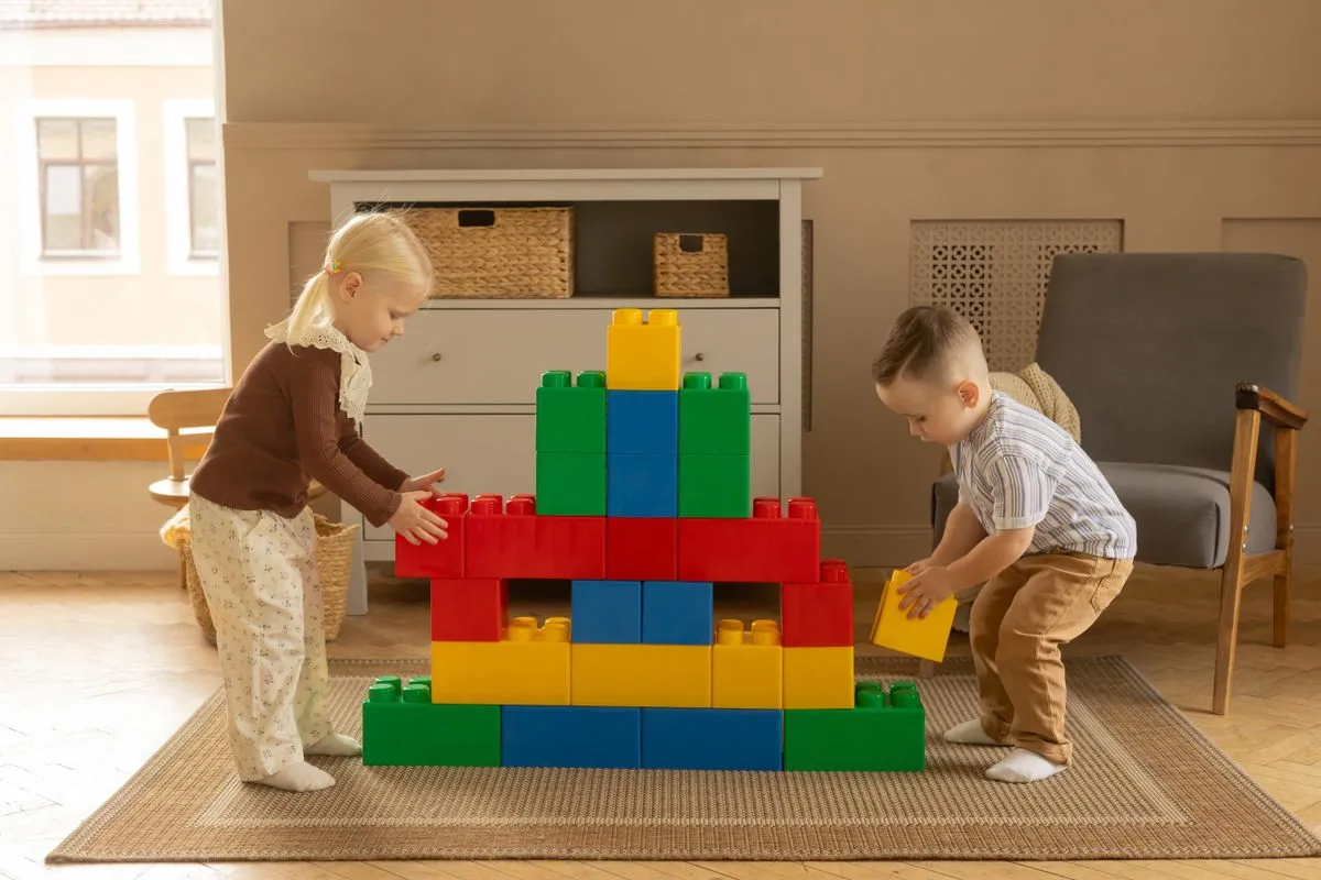 Two toddlers in a sunlit living room stacking oversized red, yellow, green and blue toy blocks into one wall together on a woven rug