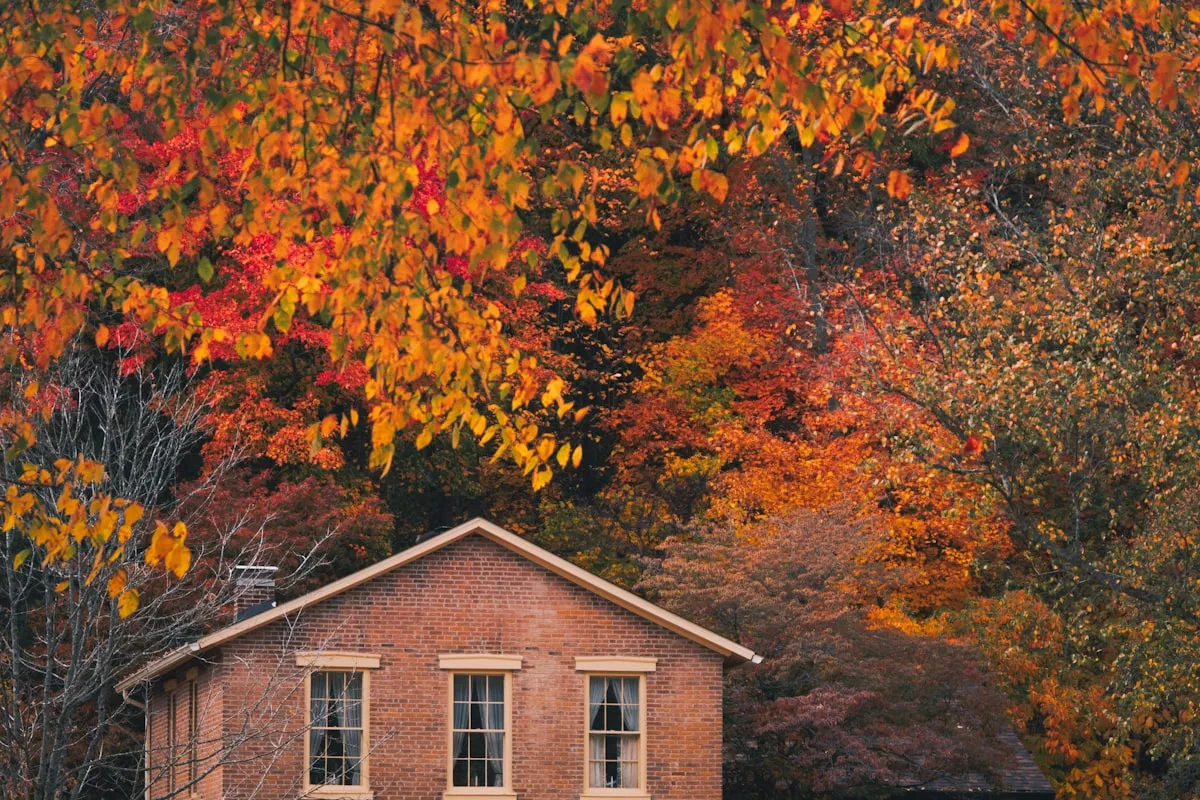 A red-brick house with a peaked roof and chimney, framed by trees in full autumn color — orange, red, and gold leaves filling the frame above the roofline.