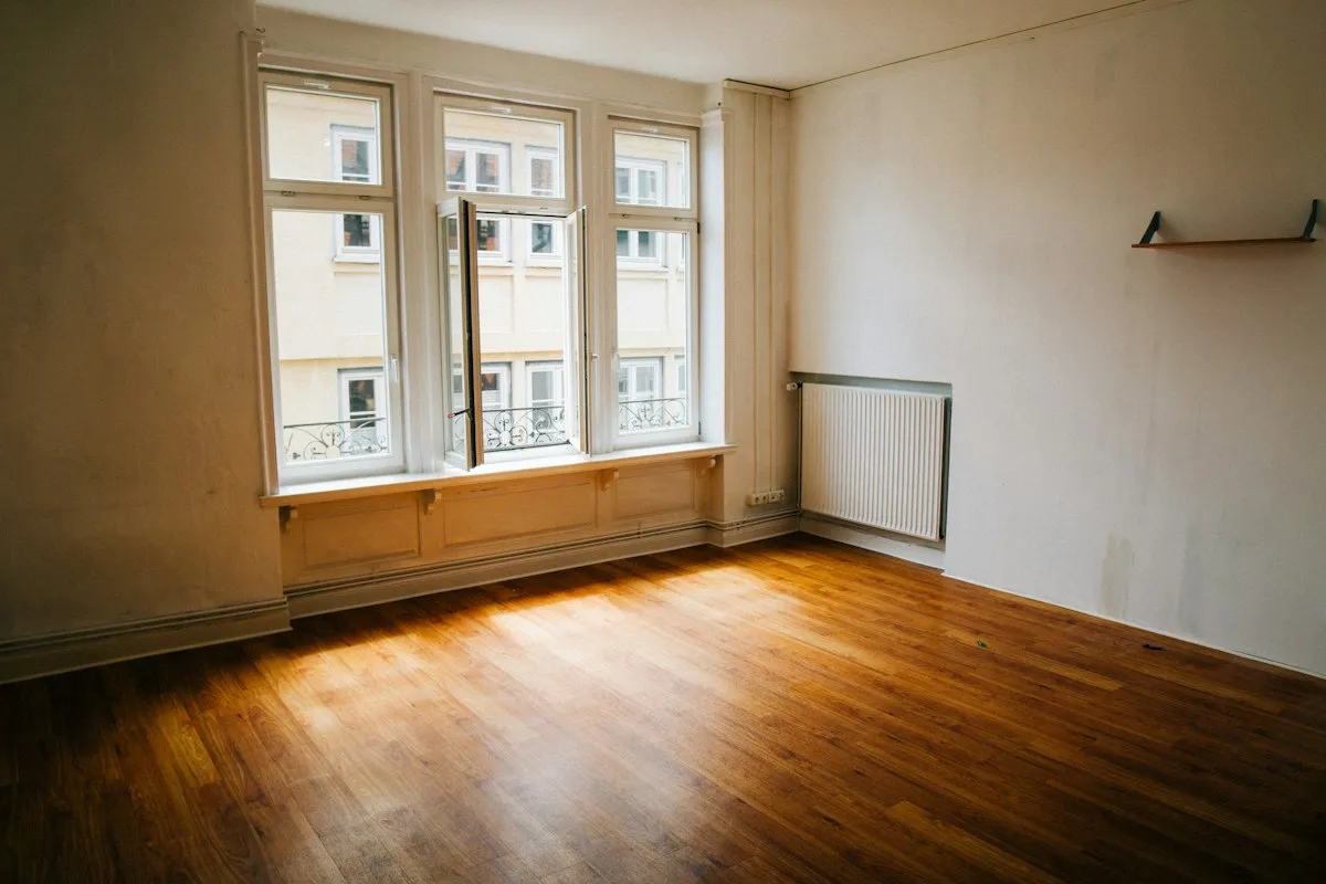 An empty apartment at turnover: sunlight pooling on bare honey-brown floorboards, three tall windows in a bay with one leaf swung open, a white panel radiator, and a shelf bracket still screwed to the wall