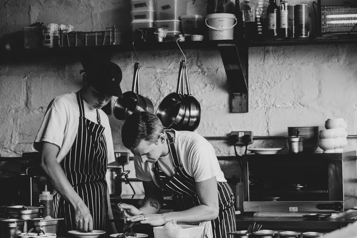 Two people in black-and-white striped aprons working side by side at a counter in a small rustic kitchen, one leaning in over plated food while the other prepares ingredients, with copper pans hanging on a whitewashed brick wall behind them