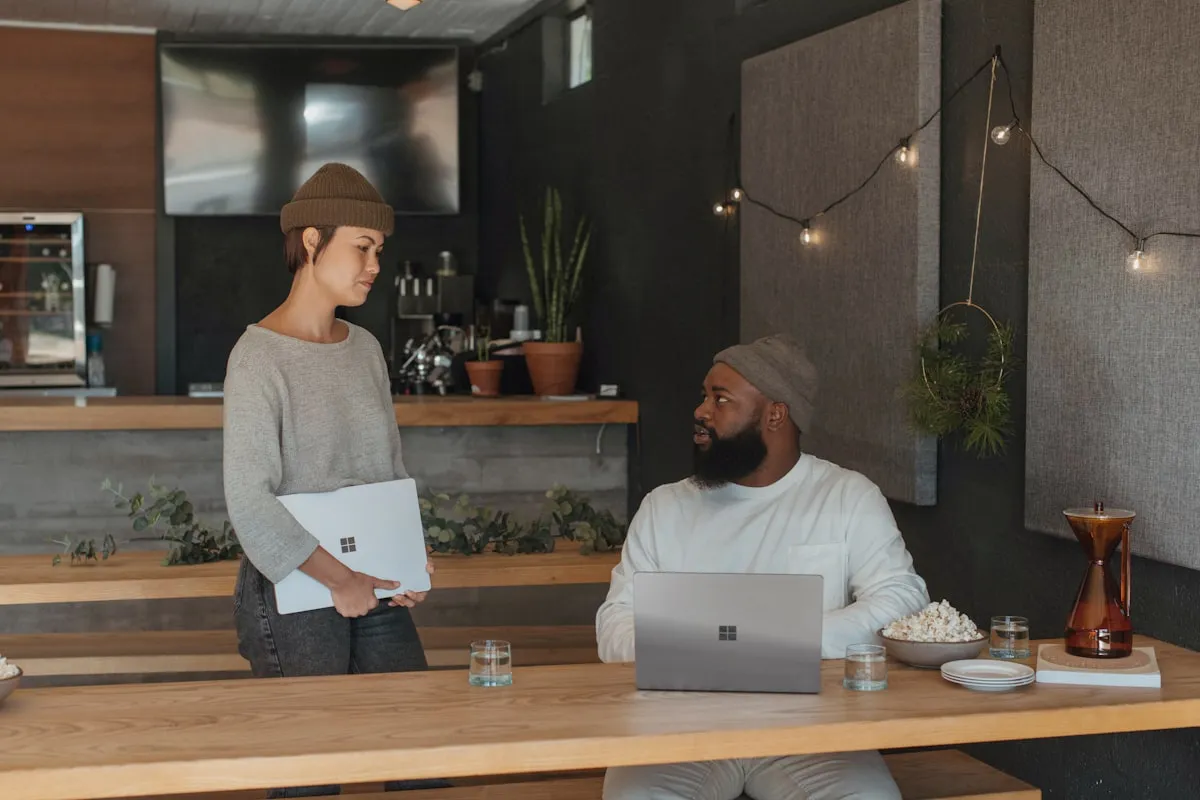 Two people in casual sweaters and knit beanies talking at a wooden counter in a warm, plant-filled cafe workspace, one standing holding a laptop and one seated with a laptop.