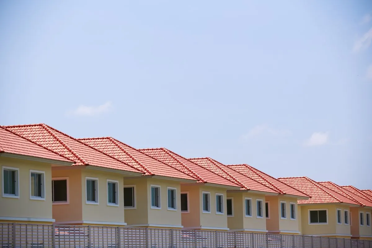 A long row of identical two-storey suburban houses with red tile roofs and pale yellow and peach walls, stretching to the right under a clear blue sky