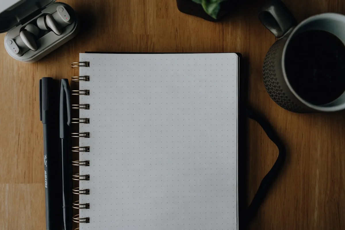An empty dot-grid notebook with two pens, wireless earbuds, and a black coffee mug arranged on a wooden desk — the aspirational productivity flat lay that productivity-routine videos open with.