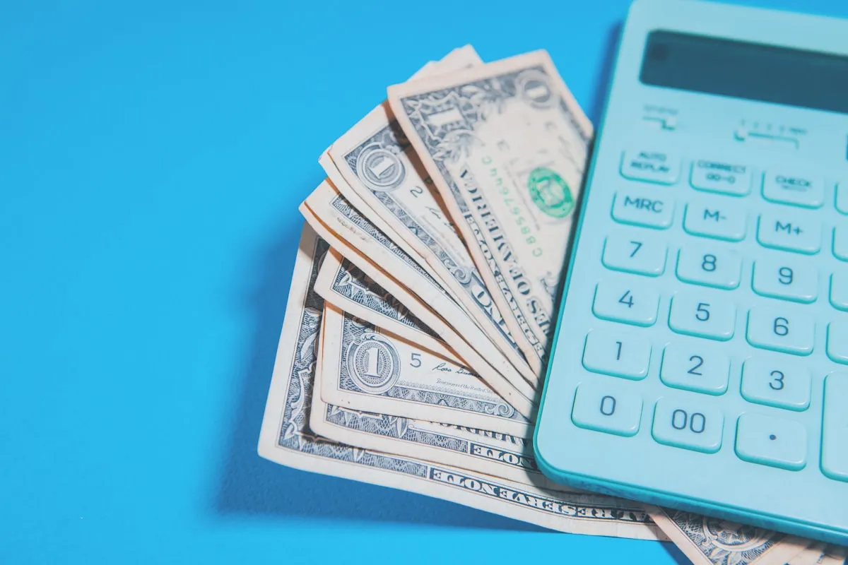 A pale teal pocket calculator resting on a fanned stack of US one-dollar bills against a bright blue background, its keypad and blank display in view