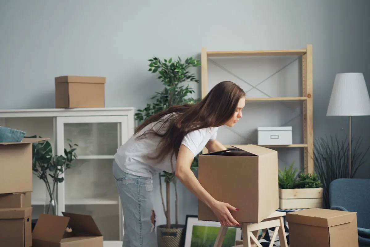 Woman in a white T-shirt and jeans bending to lift a large cardboard box in a half-packed living room with empty shelves and more boxes on the floor