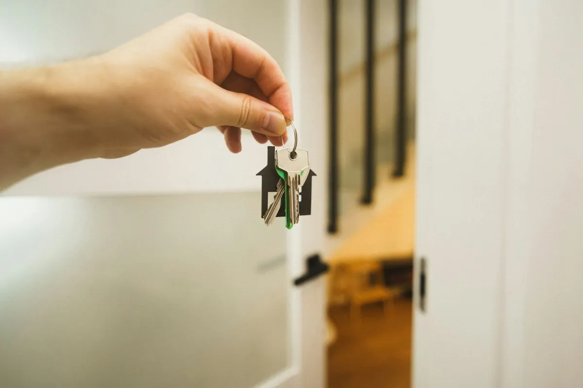 A hand holding up a small ring of house keys on a black house-shaped key tag, in front of a white interior door opening onto a sunlit room with a wooden chair