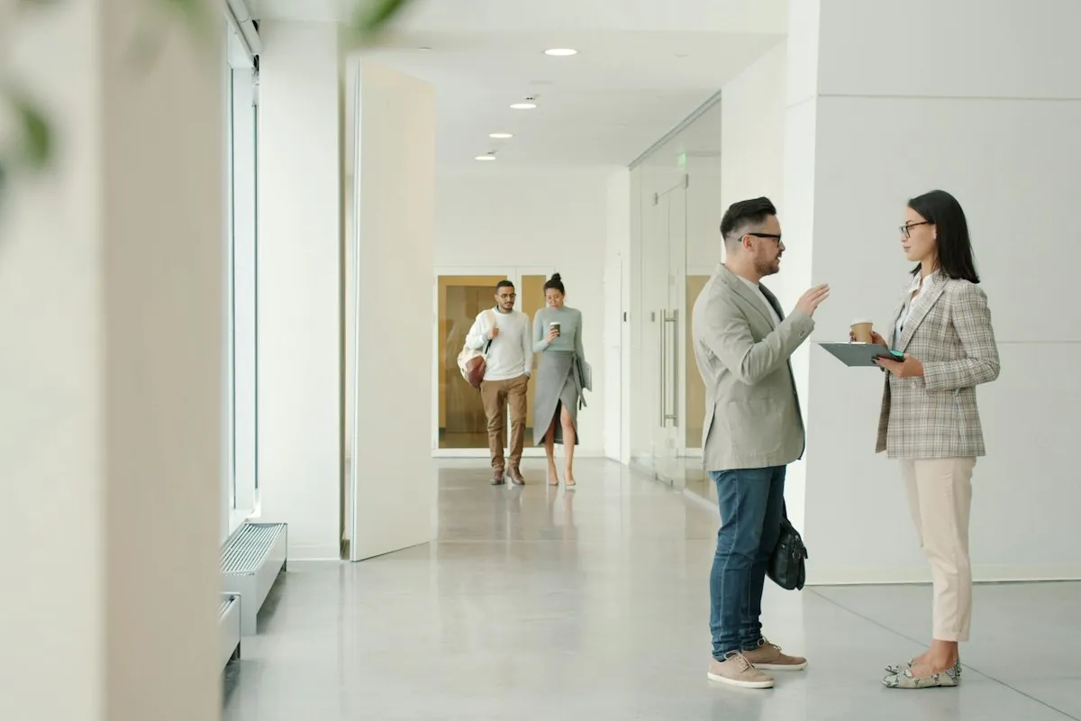 A man in a beige blazer and a woman in a checked blazer holding a clipboard and coffee cup talk face to face in a bright white office corridor while two colleagues walk toward them.