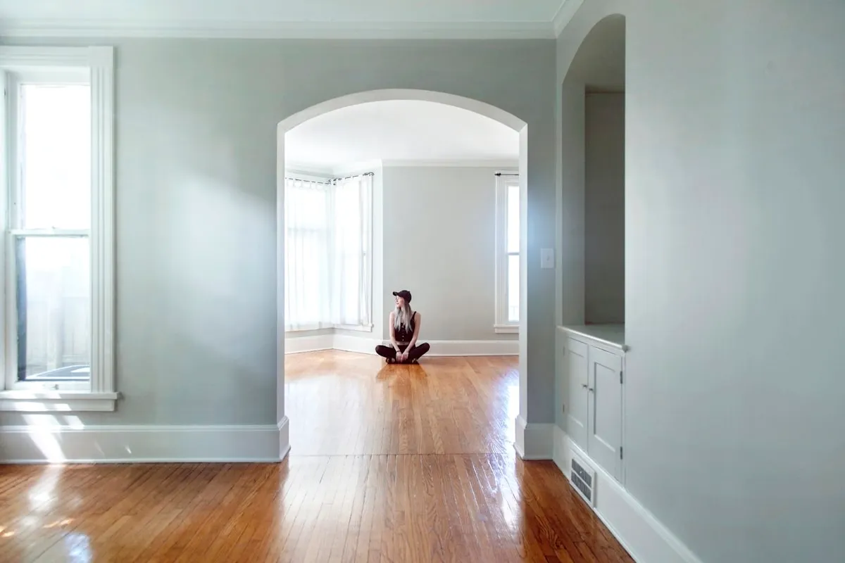 A person in a dark outfit and cap sitting cross-legged alone on the bare hardwood floor of a completely empty house, framed by a white arched doorway with sunlight coming through curtained windows