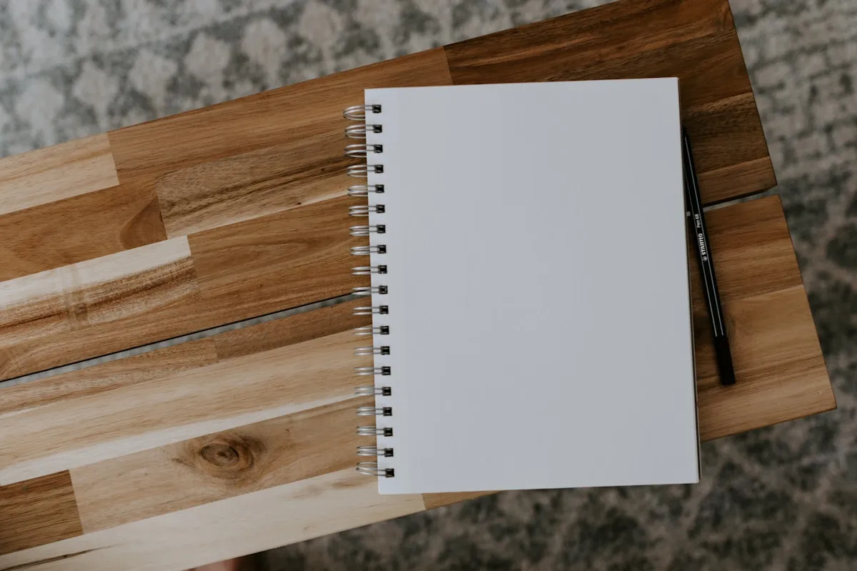 A blank white spiral-bound notebook and a black pen resting on a wood-plank board, photographed from above on a gray textured floor