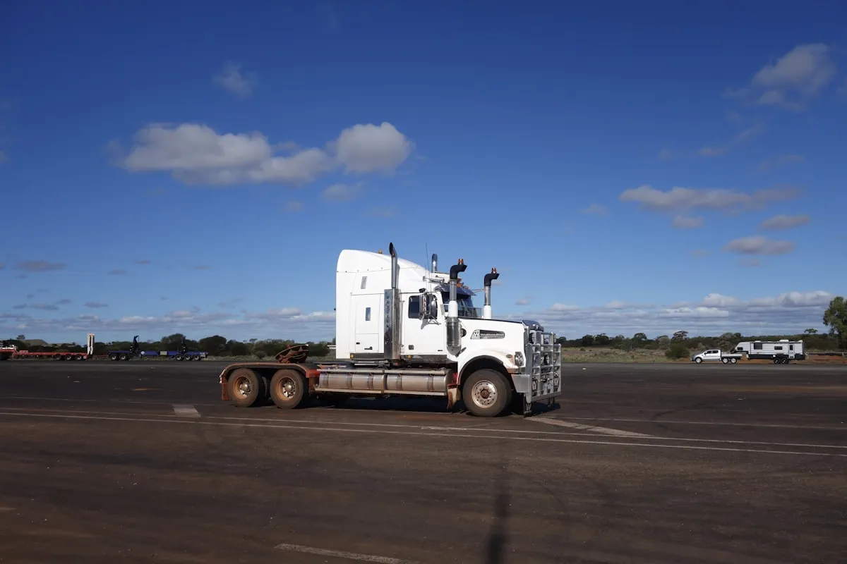A white long-nose semi tractor with no trailer attached, parked alone on a wide dusty lot under a deep blue sky, with scrub and a few distant vehicles on the horizon
