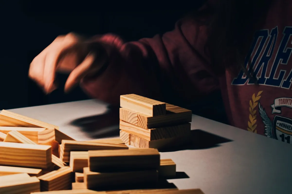A blurred hand reaching toward a small precarious stack of wooden blocks on a pale table, with dozens of loose blocks scattered in the foreground under dramatic side lighting