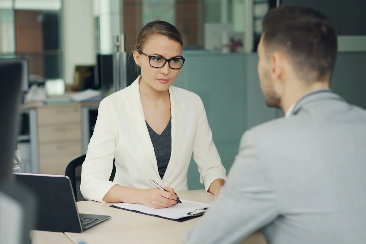 A manager in a white blazer and glasses sits across an office desk taking notes on a clipboard while interviewing a male candidate seen from behind, a laptop open beside her.